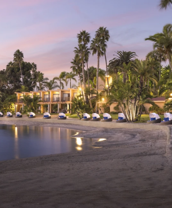 A peaceful beach at sunset with palm trees, calm water, and lounge tents along the shore, a resort line glowing softly by the sea.