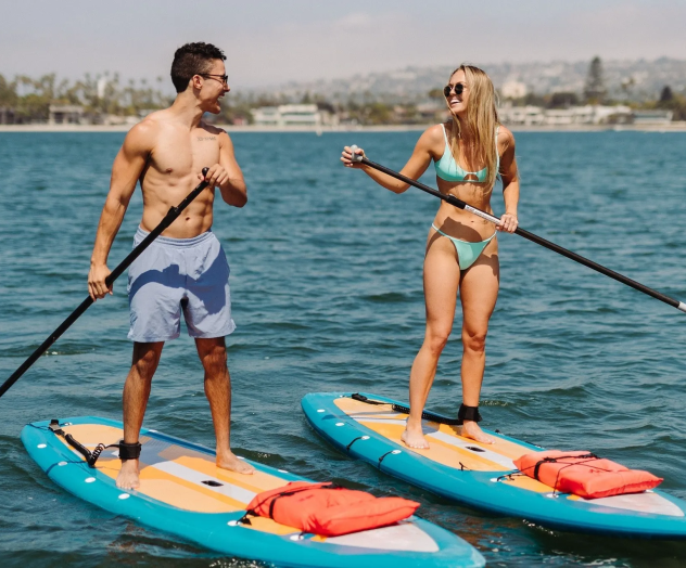 Two people stand on blue paddleboards in a sunny sea, wearing swimsuits and using long paddles while chatting and enjoying the water.