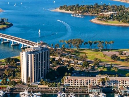 Aerial view of a coastal town with a tall building, a bridge over blue water, boats, palm trees, and a marina along the shoreline.