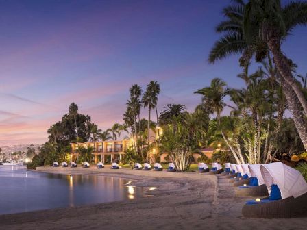 A tropical beach at sunset with calm waters, palm trees, and row of white-blue cabanas along the shore, creating a serene resort vibe.