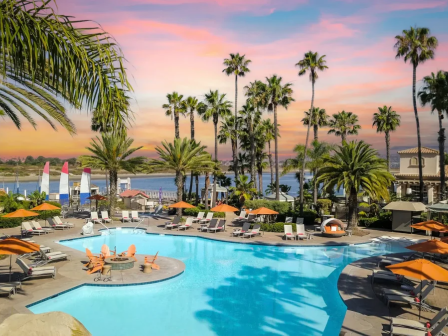 A tropical resort pool area with palm trees, lounge chairs, orange umbrellas, and a pink sunset sky over a calm ocean backdrop.