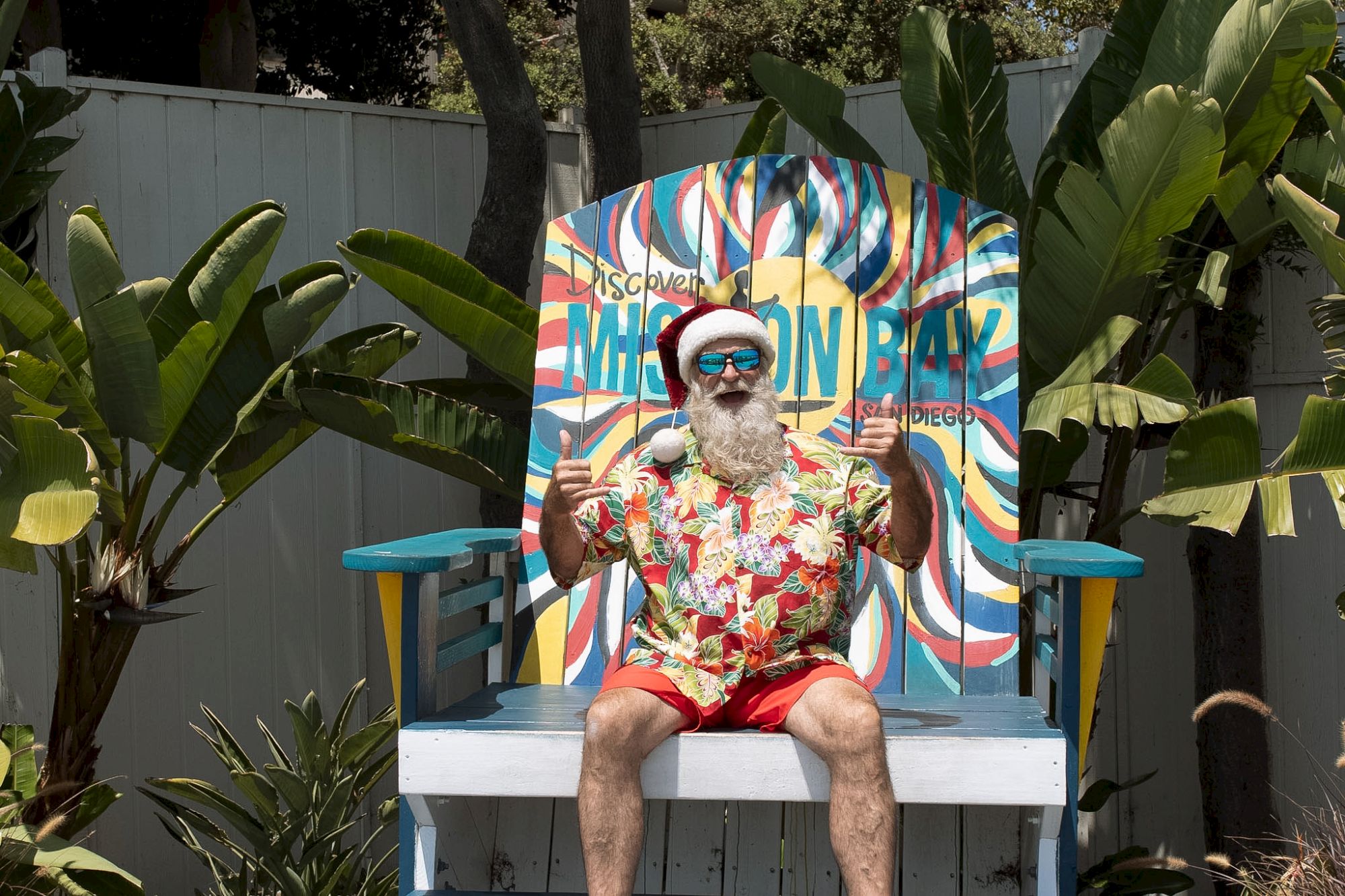 A man in a colorful Hawaiian shirt and hat sits on a large striped beach chair, with tropical plants around and a bright, psychedelic backdrop behind him.