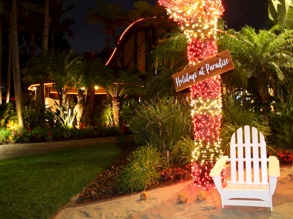 A tropical resort at night with a palm tree wrapped in orange lights, a wooden &ldquo;Welcome to Paradise&rdquo; sign, and a white Adirondack chair on sandy ground.
