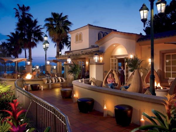 A seaside terrace at dusk with warm lighting, palm trees, lounge seating, and a villa-like restaurant overlooking the ocean, cozy and tropical.